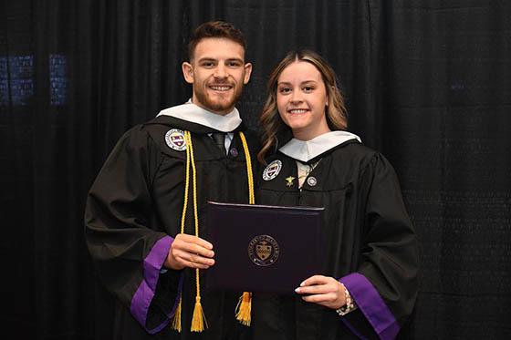 Two graduating students in robes hold up their degree and smile for a photo